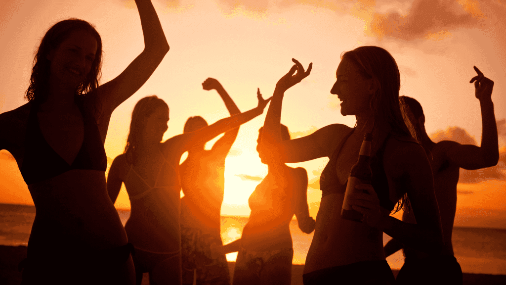 A group of people in swimwear dance and celebrate on the beach at sunset, one holding a bottle. The glowing orange sky creates vibrant silhouettes, reflecting new business opportunities in a lively coastal setting.