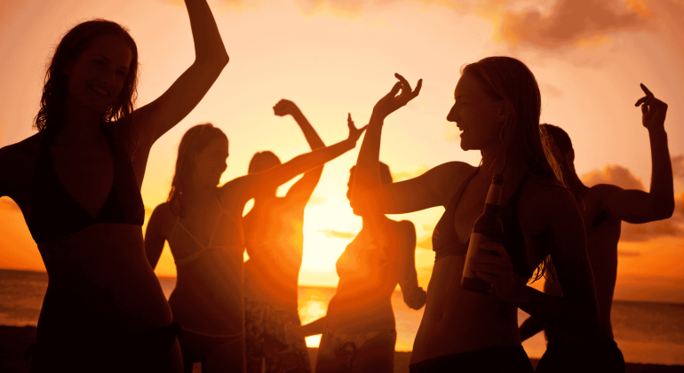 A group of people in swimwear dance and celebrate on the beach at sunset, one holding a bottle. The glowing orange sky creates vibrant silhouettes, reflecting new business opportunities in a lively coastal setting.