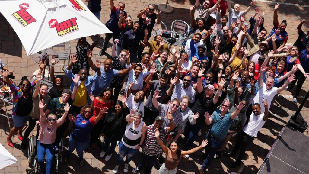 A large, diverse group of people outdoors on a patio, smiling and waving up at the camera. Some are seated, most are standing under umbrellas with HOT 1027 FM logos. It’s a bright, sunny day filled with excitement and new business opportunities.
