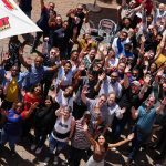A large, diverse group of people outdoors on a patio, smiling and waving up at the camera. Some are seated, most are standing under umbrellas with HOT 1027 FM logos. It’s a bright, sunny day filled with excitement and new business opportunities.