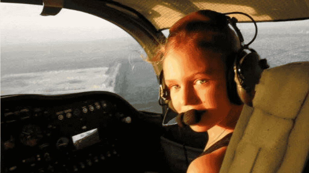 A young woman wearing a headset with a microphone sits in the cockpit of an airplane, looking back toward the camera as sunlight streams through the window, ready to explore new business opportunities.