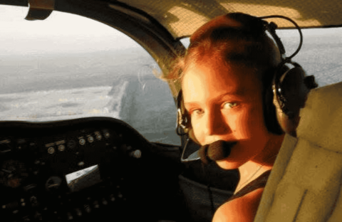 A young woman wearing a headset with a microphone sits in the cockpit of an airplane, looking back toward the camera as sunlight streams through the window, ready to explore new business opportunities.