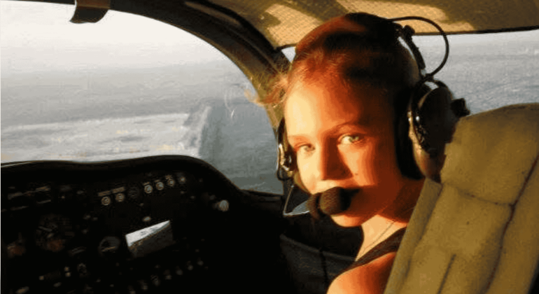 A young woman wearing a headset with a microphone sits in the cockpit of an airplane, looking back toward the camera as sunlight streams through the window, ready to explore new business opportunities.