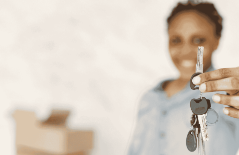 A person smiling and holding out a set of house keys, with moving boxes blurred in the background—symbolizing new beginnings and exciting business opportunities in real estate.
