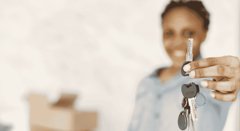 A person smiling and holding out a set of house keys, with moving boxes blurred in the background—symbolizing new beginnings and exciting business opportunities in real estate.