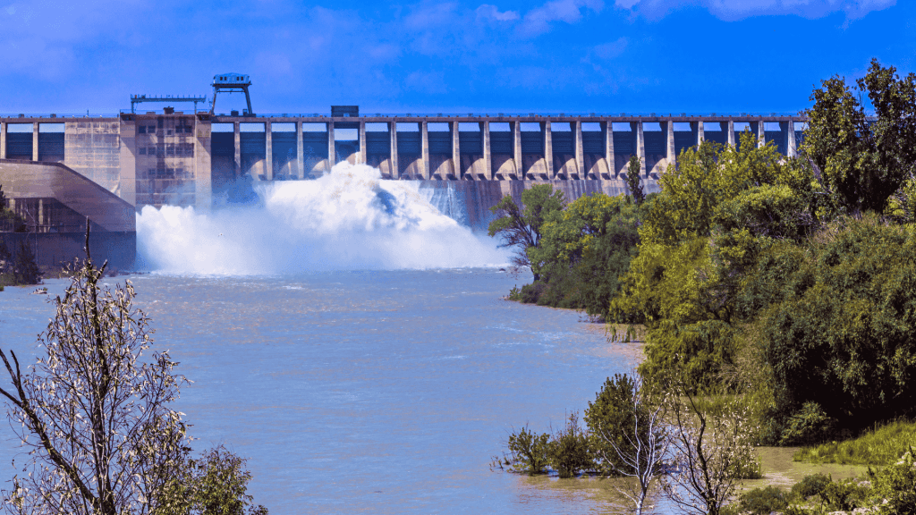 A large dam releases water into a wide river, surrounded by green trees and bushes under the bright Magaliesberg sky with scattered clouds.
