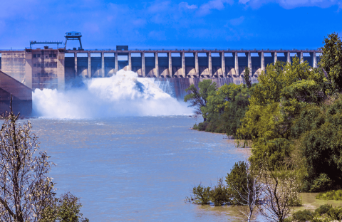 A large dam releases water into a wide river, surrounded by green trees and bushes under the bright Magaliesberg sky with scattered clouds.