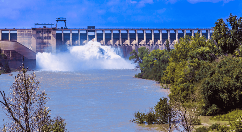 A large dam releases water into a wide river, surrounded by green trees and bushes under the bright Magaliesberg sky with scattered clouds.