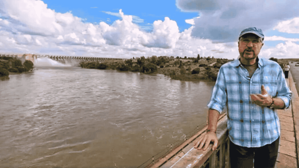 A man in a blue plaid shirt and cap stands on a bridge over a wide river near the Magaliesberg, with a dam and lush greenery in the background under a partly cloudy sky.