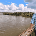 A man in a blue plaid shirt and cap stands on a bridge over a wide river near the Magaliesberg, with a dam and lush greenery in the background under a partly cloudy sky.