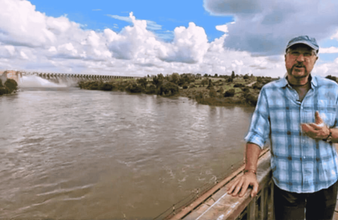 A man in a blue plaid shirt and cap stands on a bridge over a wide river near the Magaliesberg, with a dam and lush greenery in the background under a partly cloudy sky.