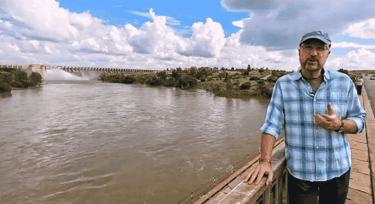 A man in a blue plaid shirt and cap stands on a bridge over a wide river near the Magaliesberg, with a dam and lush greenery in the background under a partly cloudy sky.