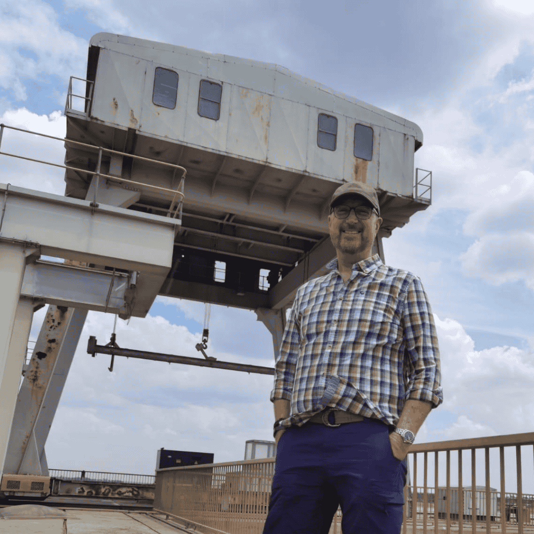 A man in a plaid shirt and cap stands smiling on an industrial platform, with large metal machinery behind him and the dramatic Magaliesberg sky overhead.