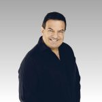 A middle-aged man with short dark hair wearing a black button-up shirt smiles at the camera against a light gray gradient background.