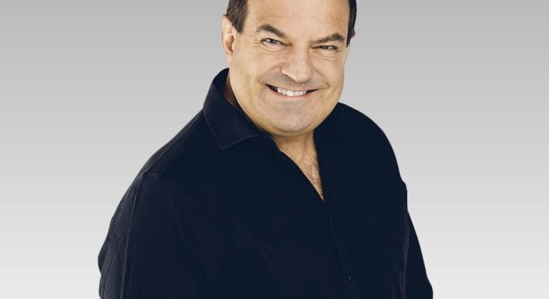 A middle-aged man with short dark hair wearing a black button-up shirt smiles at the camera against a light gray gradient background.