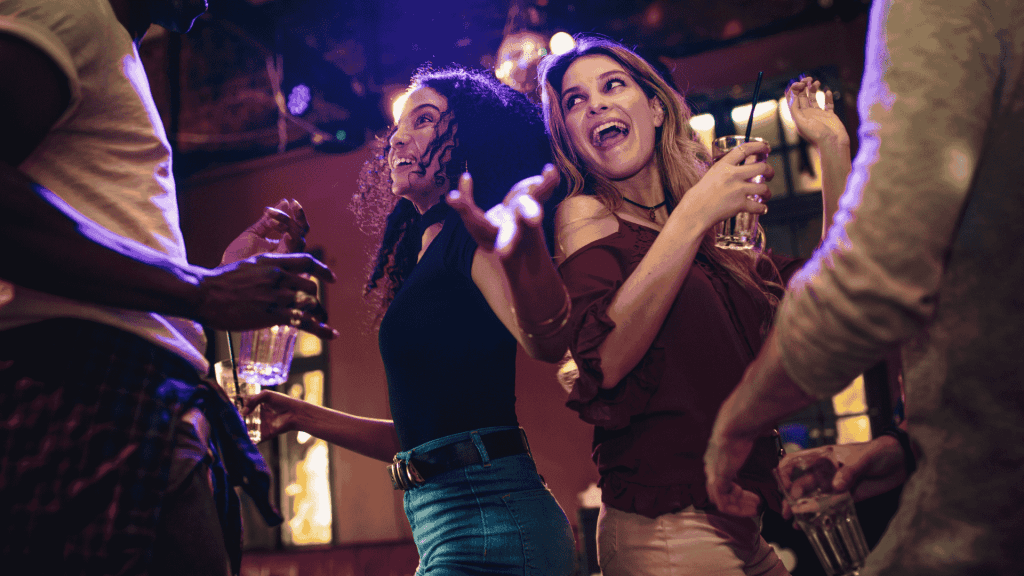 A group of people dance and smile together at a lively indoor party, holding drinks and enjoying the energetic atmosphere under colorful lights while networking for new business opportunities.