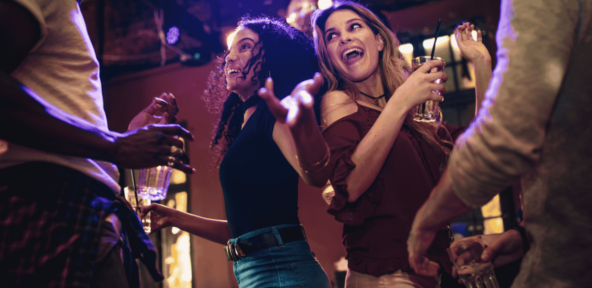 A group of people dance and smile together at a lively indoor party, holding drinks and enjoying the energetic atmosphere under colorful lights while networking for new business opportunities.