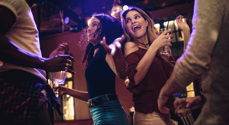 A group of people dance and smile together at a lively indoor party, holding drinks and enjoying the energetic atmosphere under colorful lights while networking for new business opportunities.