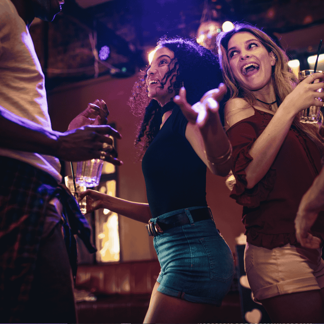 Two young women dance and laugh together at a lively indoor party, holding drinks, while a third person stands nearby. The colorful lighting and joyful expressions create a vibrant atmosphere perfect for networking or discovering new business opportunities.