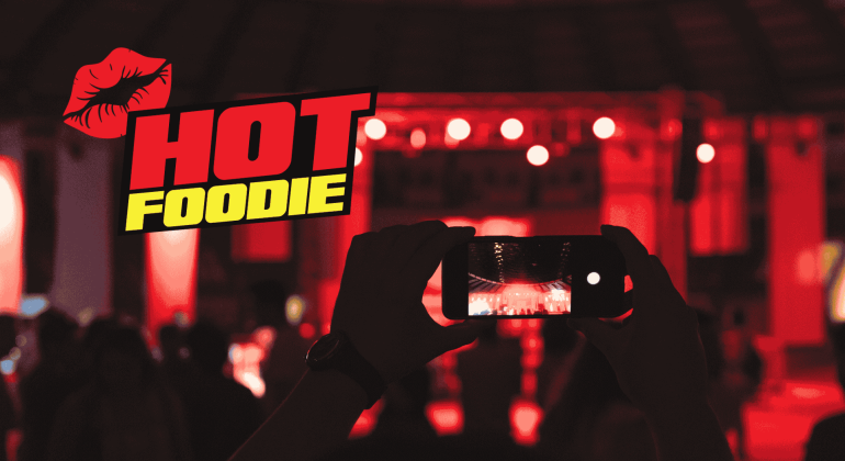 A person holds up a smartphone to take a photo at a red-lit event, with the words HOT FOODIE and a red lips graphic displayed prominently on the left side.