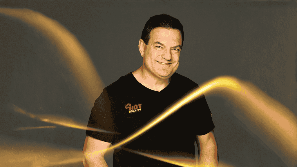 A smiling man in a black t-shirt with a HOT 102.7 FM logo stands against a dark background, illuminated by bright, swirling yellow light streaks reminiscent of Vaal Dam reflections in the foreground.