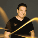 A smiling man in a black t-shirt with a HOT 102.7 FM logo stands against a dark background, illuminated by bright, swirling yellow light streaks reminiscent of Vaal Dam reflections in the foreground.