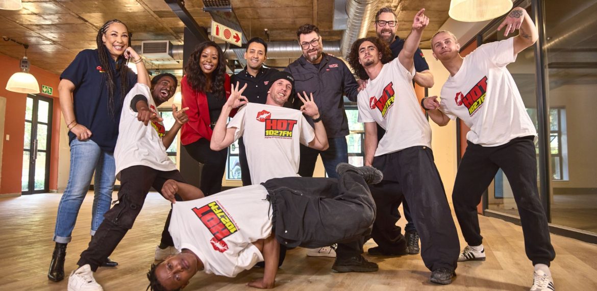 A group of nine people, some standing and some posing energetically—one even breakdancing—smile and gesture playfully in a modern indoor setting with wooden floors and large windows, capturing the lively spirit of Hot 102.7fm.