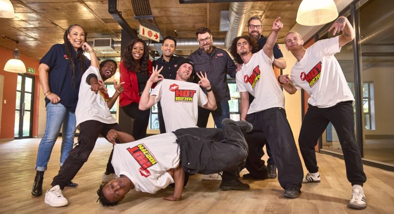 A group of nine people, some standing and some posing energetically—one even breakdancing—smile and gesture playfully in a modern indoor setting with wooden floors and large windows, capturing the lively spirit of Hot 102.7fm.