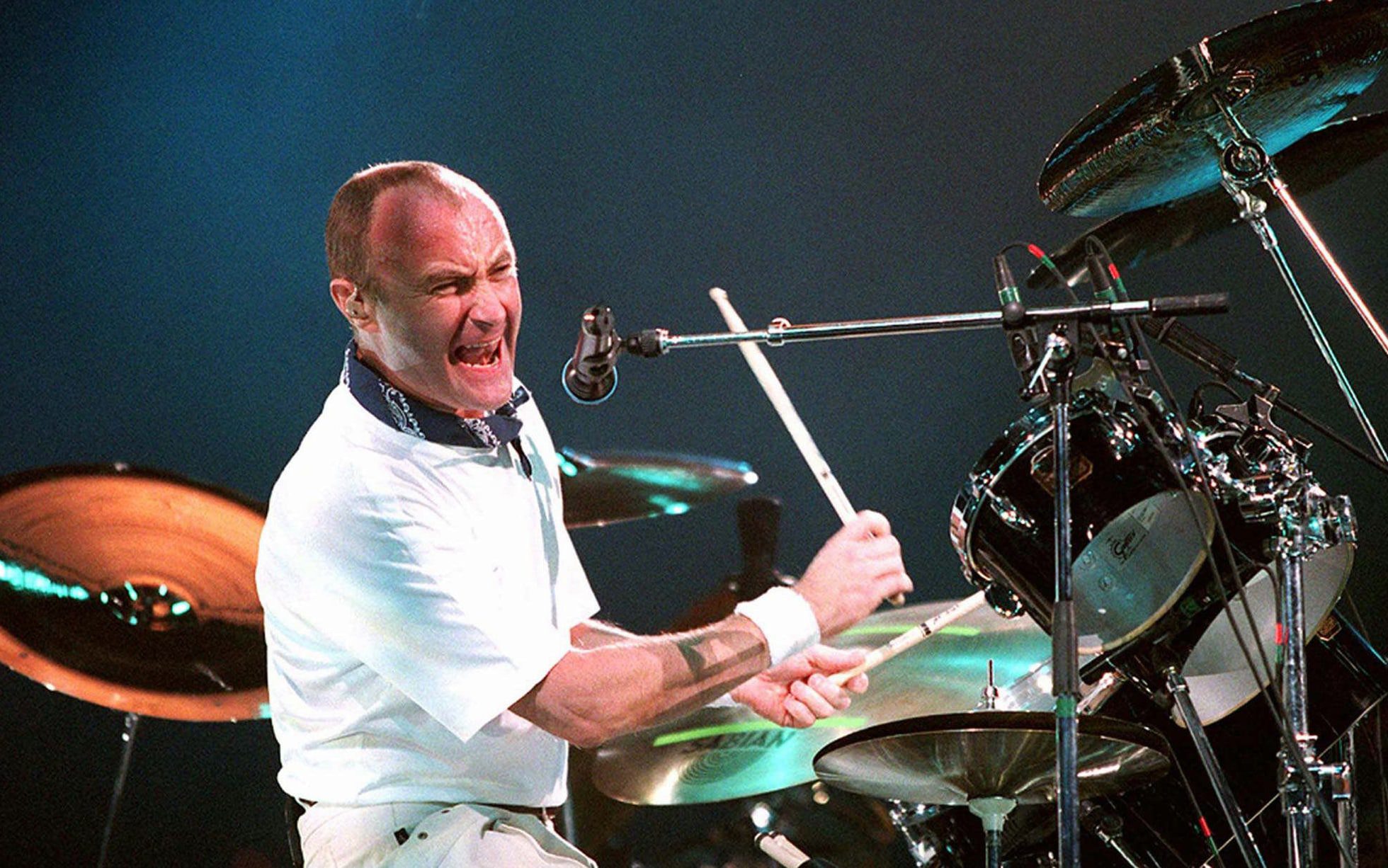 A drummer passionately plays a Matric drum set on stage, mid-performance. He is wearing a white outfit and is captured with an intense expression, holding drumsticks, surrounded by cymbals and drums.