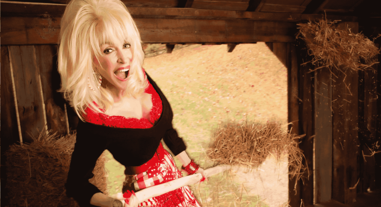 A cheerful woman with blonde hair, dressed in a red and black outfit, stands inside a rustic wooden barn—her Matric sash draped proudly as she holds a pitchfork, surrounded by straw.