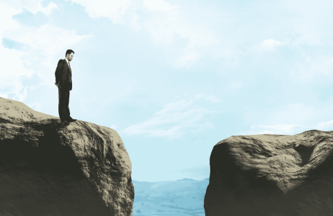 A man in a suit stands on the edge of a cliff, looking across a wide gap toward another rocky ledge under a blue sky with clouds, contemplating new business opportunities.