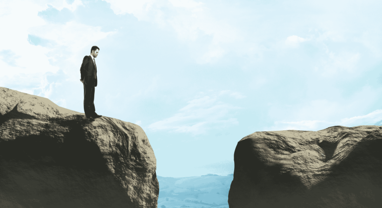 A man in a suit stands on the edge of a cliff, looking across a wide gap toward another rocky ledge under a blue sky with clouds, contemplating new business opportunities.