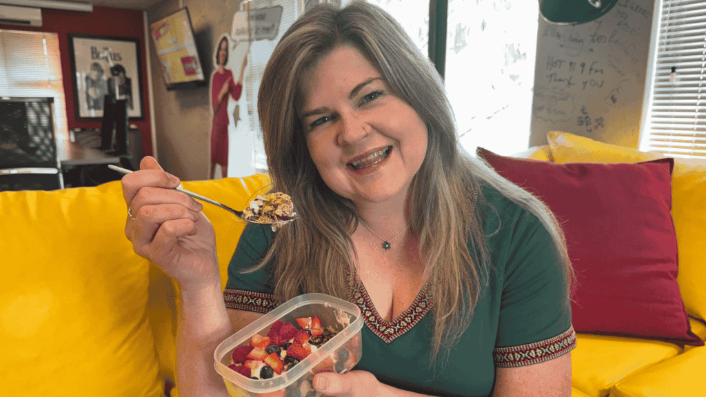 A smiling woman with long hair sits on a yellow couch, holding a container of fruit and a spoon. Wearing a green top, she enjoys her snack in a cozy, colorful room where cover versions of songs play softly in the background.