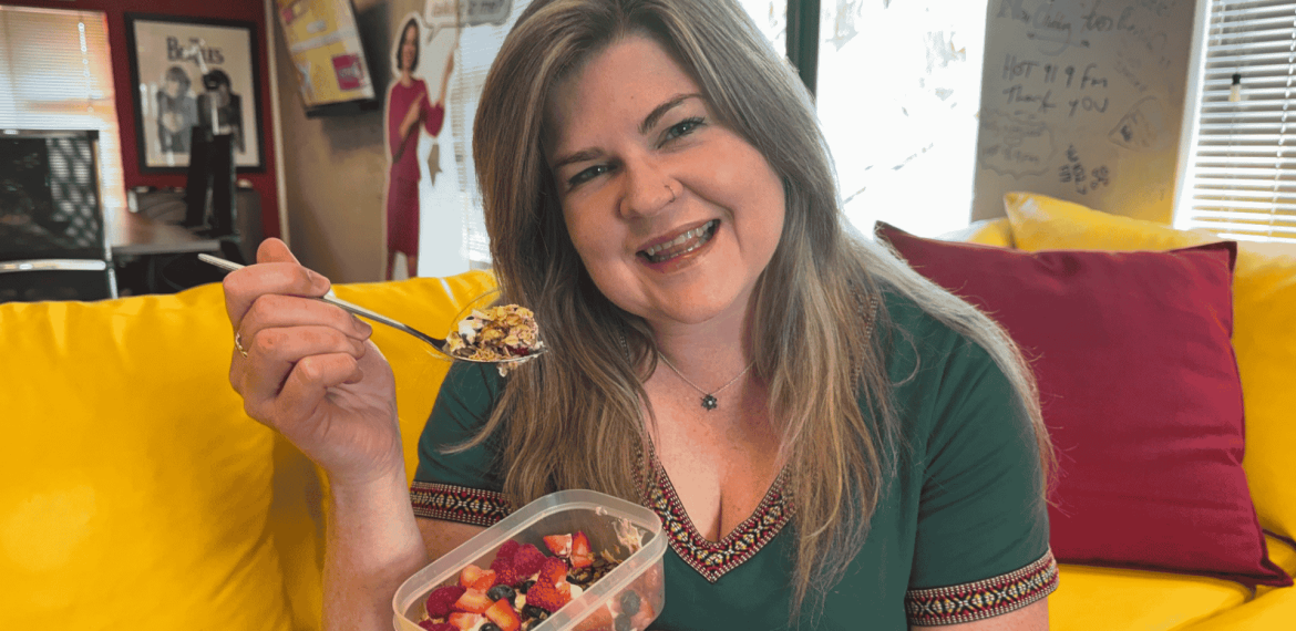 A smiling woman with long hair sits on a yellow couch, holding a container of fruit and a spoon. Wearing a green top, she enjoys her snack in a cozy, colorful room where cover versions of songs play softly in the background.
