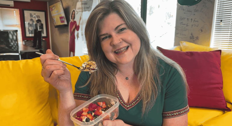 A smiling woman with long hair sits on a yellow couch, holding a container of fruit and a spoon. Wearing a green top, she enjoys her snack in a cozy, colorful room where cover versions of songs play softly in the background.