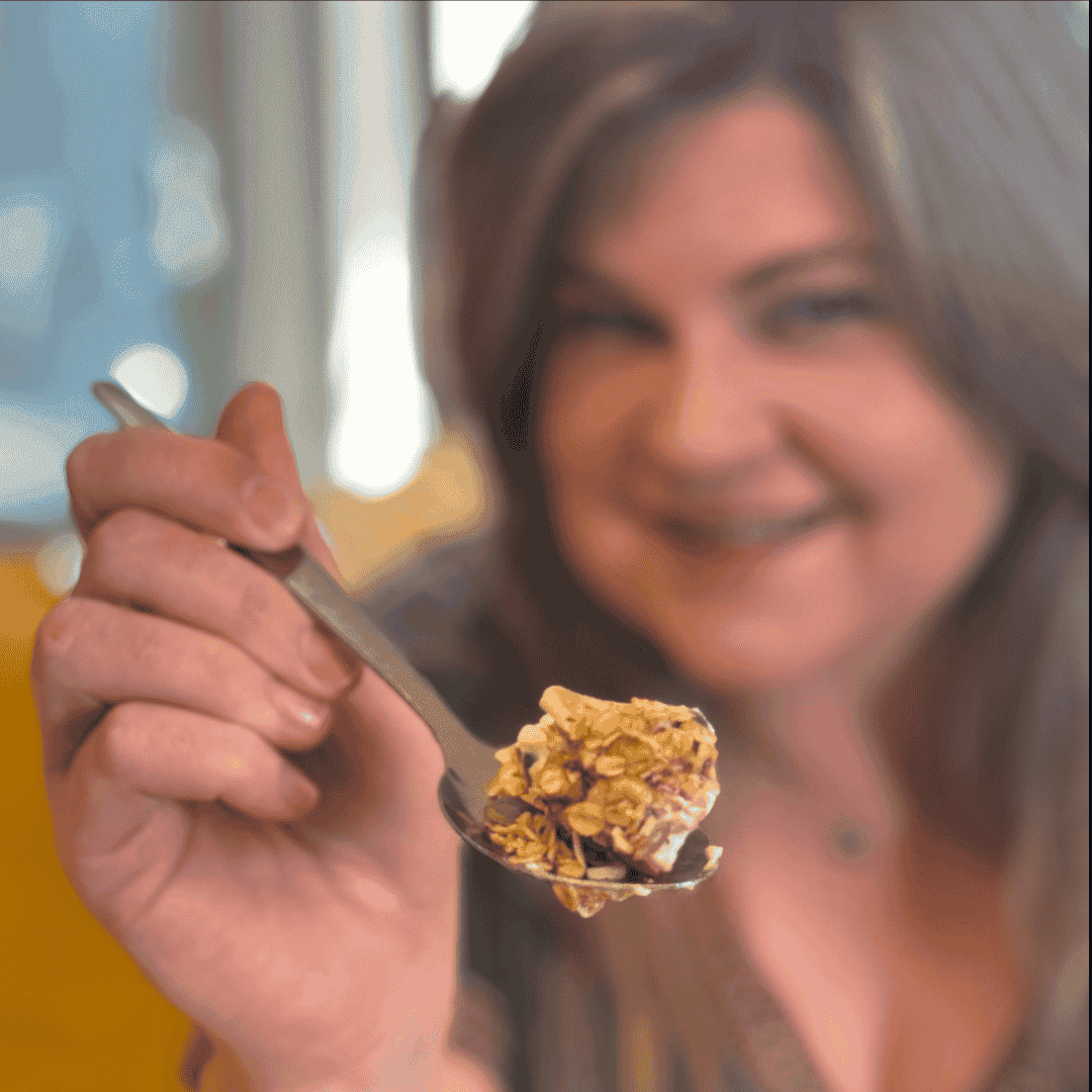 A woman smiles and holds a spoonful of yogurt topped with granola and berries toward the camera, her face slightly out of focus in the background, creating a scene that feels as inviting as creative cover versions of classic favorites.
