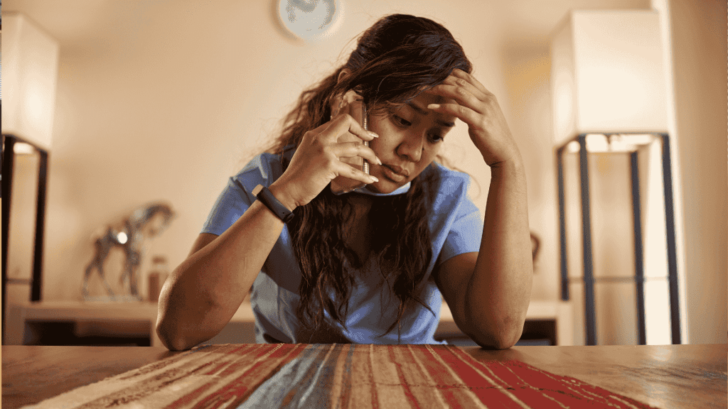 A woman in scrubs sits at a table, looking stressed while talking on the phone about business opportunities, her hand resting on her forehead.