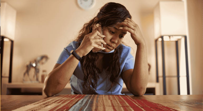 A woman in scrubs sits at a table, looking stressed while talking on the phone about business opportunities, her hand resting on her forehead.
