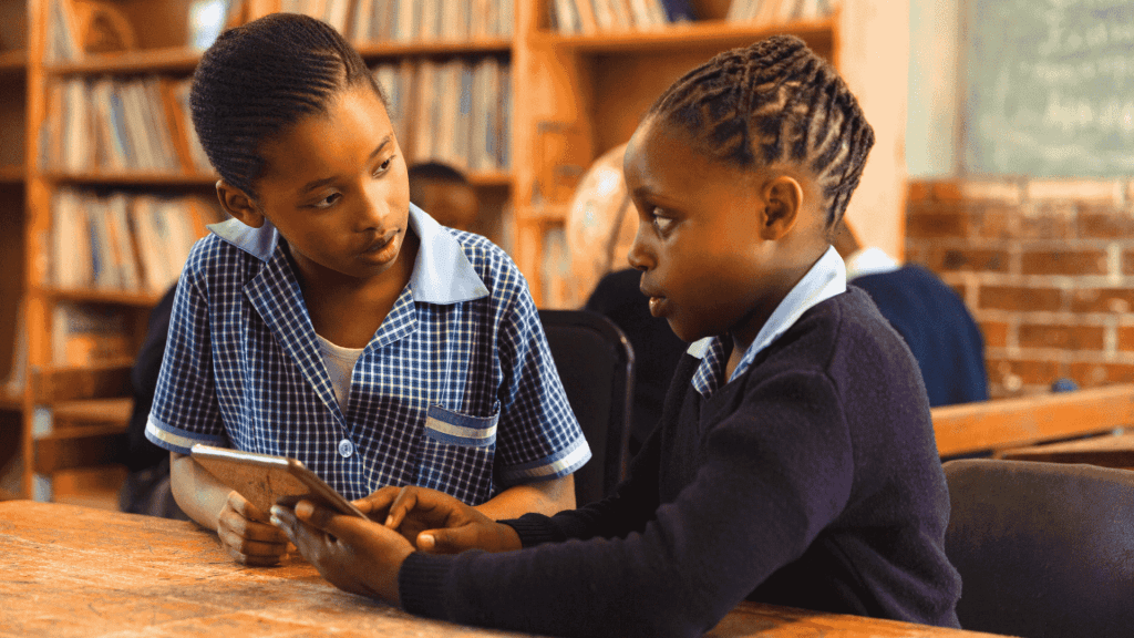 Two schoolchildren in uniform sit at a wooden table in a library, engaged in conversation. One holds a tablet—perhaps reviewing Matric notes—while the other listens attentively. Bookshelves and a chalkboard are visible in the background.