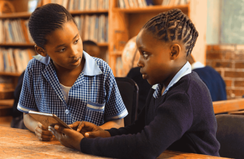 Two schoolchildren in uniform sit at a wooden table in a library, engaged in conversation. One holds a tablet—perhaps reviewing Matric notes—while the other listens attentively. Bookshelves and a chalkboard are visible in the background.