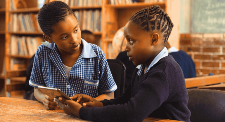 Two schoolchildren in uniform sit at a wooden table in a library, engaged in conversation. One holds a tablet—perhaps reviewing Matric notes—while the other listens attentively. Bookshelves and a chalkboard are visible in the background.