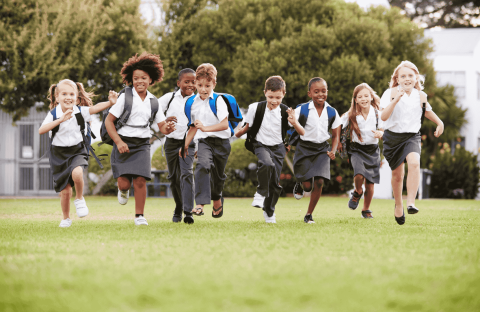 A diverse group of young children in school uniforms run happily across a grassy field, carrying backpacks, with trees and buildings—symbolizing future business opportunities—in the background.