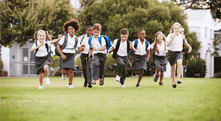 A diverse group of young children in school uniforms run happily across a grassy field, carrying backpacks, with trees and buildings—symbolizing future business opportunities—in the background.