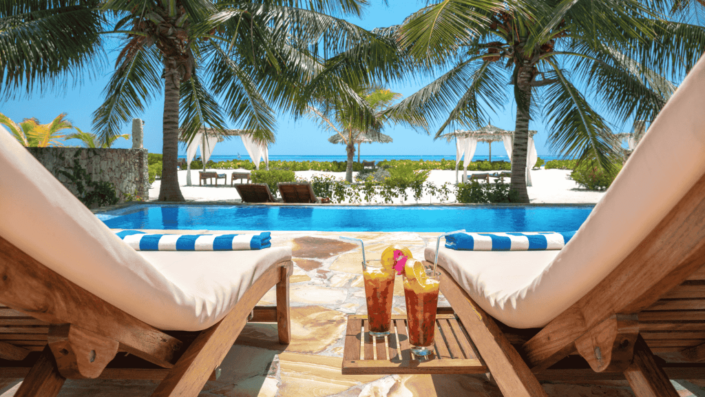 Two lounge chairs by a pool overlook a tropical beach with palm trees and turquoise ocean. Matric exams behind you, sip iced drinks with fruit garnish under sunny skies, relaxing by the white canopies and gentle waves.