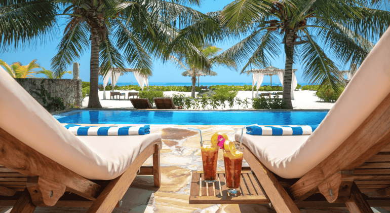 Two lounge chairs by a pool overlook a tropical beach with palm trees and turquoise ocean. Matric exams behind you, sip iced drinks with fruit garnish under sunny skies, relaxing by the white canopies and gentle waves.