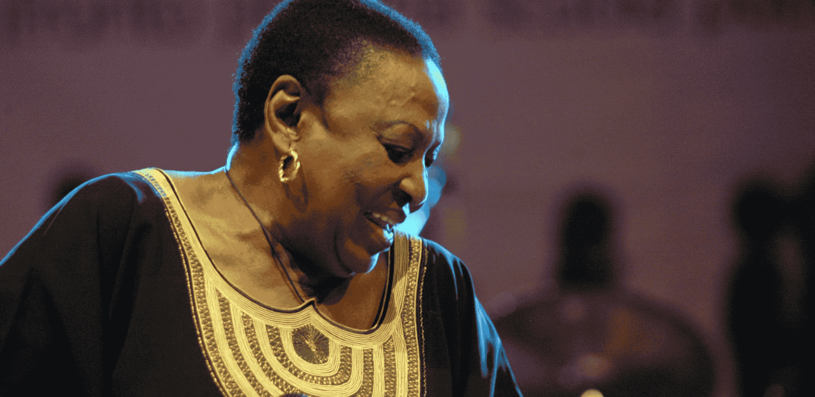 A woman wearing a black and gold traditional outfit smiles while holding a microphone on stage in Yeoville, with blurred figures and a purple-lit background behind her.