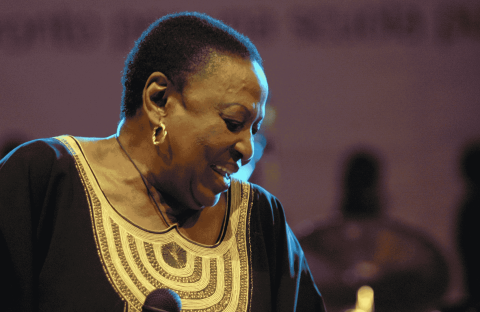 A woman wearing a black and gold traditional outfit smiles while holding a microphone on stage in Yeoville, with blurred figures and a purple-lit background behind her.