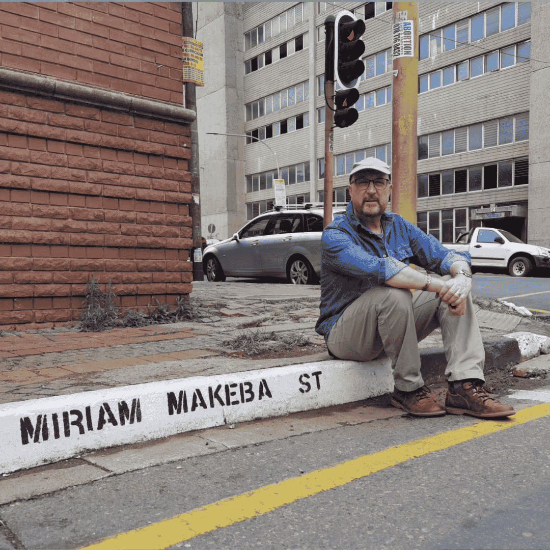 A man in casual clothes and a cap sits on a curb labeled MIRIAM MAKEBA ST in Yeoville, beside a city street with cars, buildings, and a traffic light in the background.