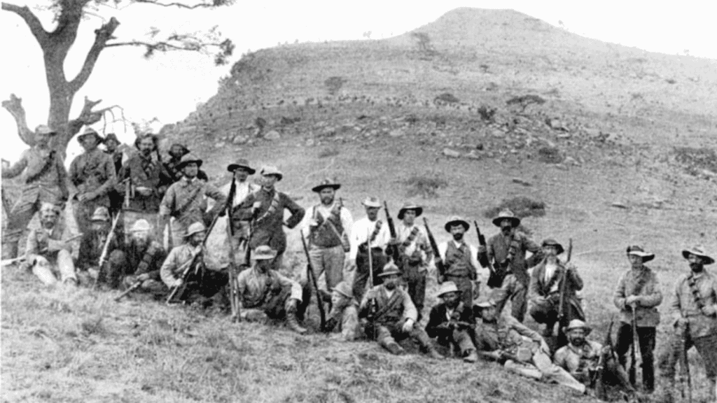 A group of men in hats and old-fashioned clothing pose with rifles on a grassy hillside near Yeoville; a lone tree stands to the left and a rugged hill rises in the background. The photo appears to be historical.
