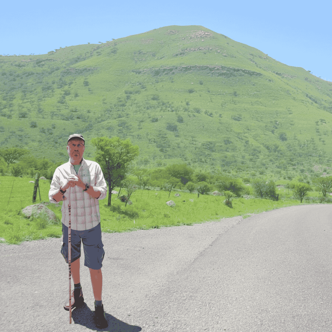 A man wearing a hat, glasses, and hiking clothes stands on the side of a paved road in Yeoville, holding a walking stick, with a green grassy hill and scattered trees in the background under a clear blue sky.